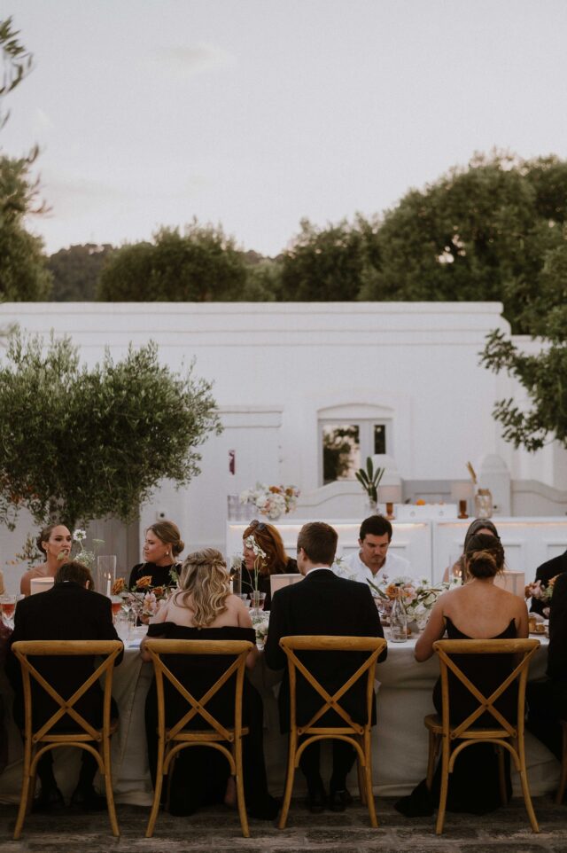 Un gruppo di persone eleganti sedute a un tavolo lungo durante un evento all'aperto, circondati da una scenografia raffinata