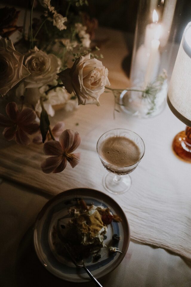 Un dessert su un piatto accanto a un bicchiere di cocktail, circondato da decorazioni floreali e candele